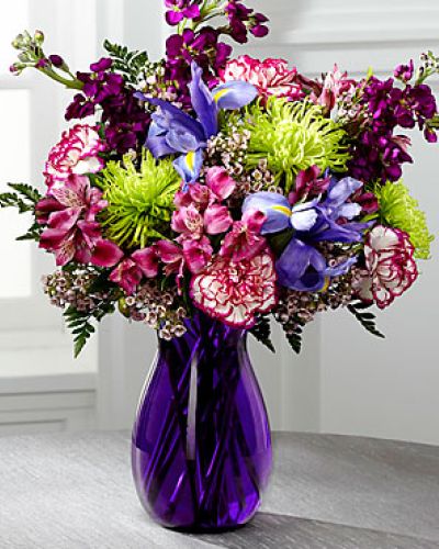 Blooming with rich color and undeniable texture, this stunning bouquet expresses love and gratitude with each perfect flower. Blue Iris, fragrant purple gilly flower, purple Peruvian Lilies, purple bi-color carnations, green spider chrysanthemums, clusters of pink wax flower, and lush greens create a fascinating display presented in a deep purple glass vase with modern lines to create a sophisticated look.